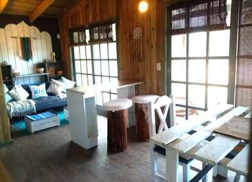 This cozy rustic room features wood-paneled walls, large windows, a white pallet dining table, log stools, a couch with cushions, and ample natural light streaming in.