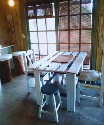 A rustic, cozy dining area with wooden furniture, pallet table, assorted chairs, natural light from window, bamboo blinds, and log stools create a warm, inviting atmosphere.