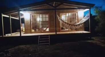 A cozy wooden cabin is warmly lit at night, featuring a spacious porch with a hammock, large windows, and inviting steps leading up to the entrance.
