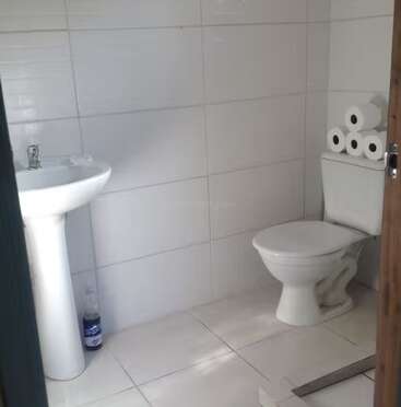 This image shows a simple, clean bathroom with white tiled walls and floors, a pedestal sink, a toilet, several rolls of toilet paper, and a soap bottle.
