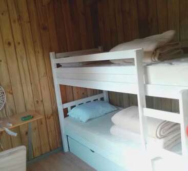 This image shows a cozy wooden room with a white bunk bed, neatly arranged blankets and pillows, a small table, a fan, and wooden walls.