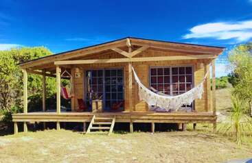 A charming wooden cabin sits on stilts with a cozy front porch, two hammocks, large windows, surrounded by greenery, under a vibrant blue sky.