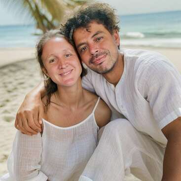 A couple sits closely together on a sandy beach, smiling warmly. Both wear white clothing, with the ocean and palm trees visible in the serene background.