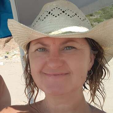 A imagem mostra uma mulher usando um chapéu de caubói de palha e sorrindo para a câmera, com uma praia e um terreno rochoso ao fundo.
