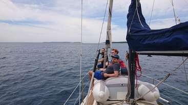Three people relax at the bow of a sailboat, enjoying calm seas and clear skies. Land is visible in the distance, completing this peaceful maritime scene.