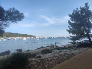 A tranquil seaside scene features several boats anchored in calm blue waters, people relaxing near the shore, scattered rocks, and lush green trees under a clear sky.