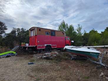 A bright red converted truck serves as a tiny house in a natural setting. Nearby, a green boat rests on a trailer amid scattered wooden planks.