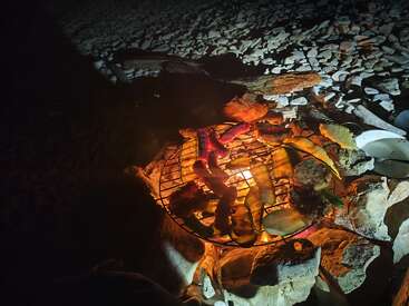 A nighttime outdoor barbecue scene shows vegetables and meats grilling over an open fire, surrounded by rocks, with a rocky ground illuminated by the firelight.