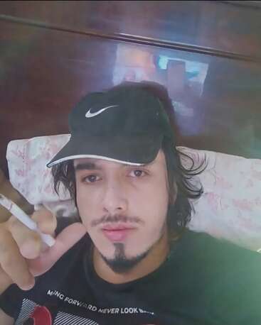Ein junger Mann mit Gesichtsbehaarung und langen Haaren entspannt sich auf einem Bett, trägt eine schwarze Nike-Mütze, hält eine Zigarette in der Hand und schaut direkt in die Kamera.