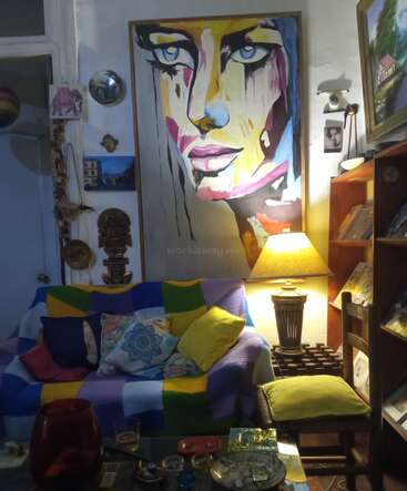 Ein gemütliches Wohnzimmer mit einem farbenfrohen Patchwork-Sofa, lebendigen Kissen, einem auffälligen abstrakten Porträt, warmer Lampenbeleuchtung, Bücherregalen und eklektischer Wandkunst und Dekoration.