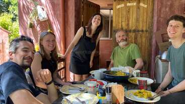 Cinco personas están sentadas alrededor de una mesa al aire libre, disfrutando de una comida juntas. Todas sonríen y parecen felices. El entorno es luminoso y acogedor.