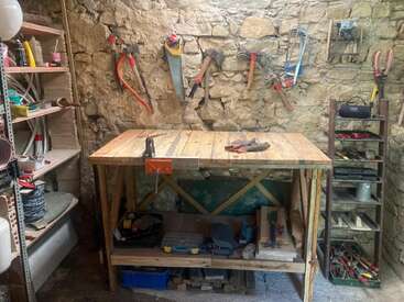 Esta imagen muestra un taller rústico con un banco de trabajo de madera, varias herramientas manuales en una pared de piedra, estantes llenos de suministros y equipos organizados por todas partes.