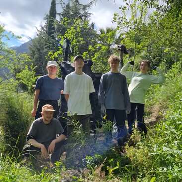 Cinco jóvenes posan juntos en un entorno exterior exuberante y verde. Visten ropa informal y gorras, rodeados de árboles y follaje, disfrutando de la belleza de la naturaleza.
