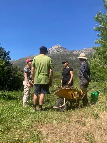 Four people stand outdoors on grass, surrounded by greenery and mountains with a clear blue sky. A wheelbarrow and watering can sit nearby, suggesting gardening work.