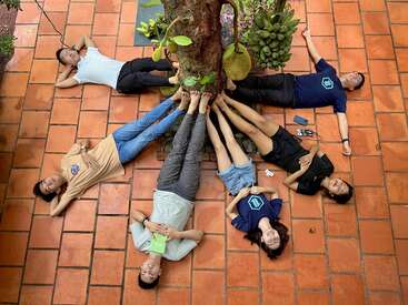 L'image représente six personnes allongées sur le sol en formation circulaire autour d'un tronc d'arbre, la tête dirigée vers le centre.