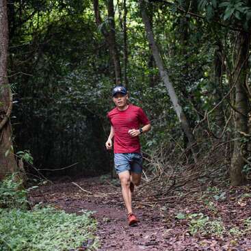 Un homme vêtu d'une chemise rouge et d'un short bleu court sur un sentier dans les bois, entouré d'arbres et de verdure, en portant une montre à son poignet gauche.