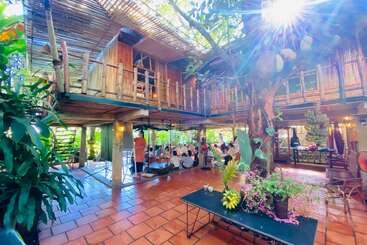 Cette image montre un restaurant rustique en plein air avec une architecture en bambou, des plantes vertes luxuriantes, la lumière du soleil, un grand arbre central et des gens qui dînent ensemble dans la bonne humeur.