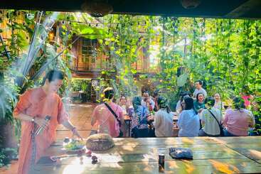Un groupe de personnes est assis à une longue table dans une cour de jardin luxuriante et ensoleillée. Une femme se tient à proximité, préparant de la nourriture sur une table en bois.