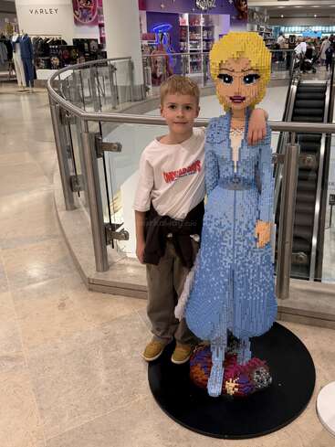 Um menino está ao lado de uma grande escultura de LEGO de uma mulher com um vestido azul dentro de um shopping center, sorrindo para a câmera.