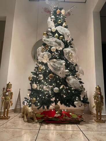 Un arbre de Noël magnifiquement décoré avec des ornements dorés et argentés, un ruban en maille blanche, deux personnages de casse-noisettes dorés, un animal doré et une jupe de sapin rouge festive.