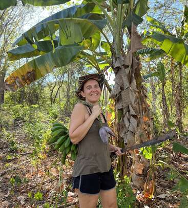 Una mujer sonriente junto a un platanero, con un machete en la mano y plátanos recién cosechados. Lleva sombrero, camiseta de tirantes y pantalones cortos en un entorno exuberante.
