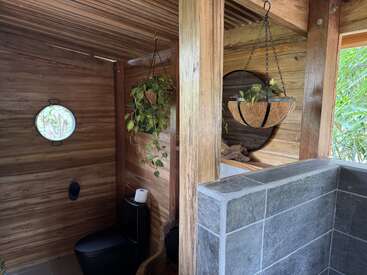 Esta imagen muestra un baño rústico con paredes de madera, un inodoro negro, una ventana redonda, plantas colgantes, un espejo redondo y una mampara de ducha parcial con azulejos de piedra. La luz natural entra a raudales.