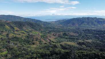 Cette image montre des collines et des montagnes verdoyantes avec des forêts denses. Au loin, on aperçoit un lac ou une mer d'un bleu paisible sous un ciel partiellement nuageux.