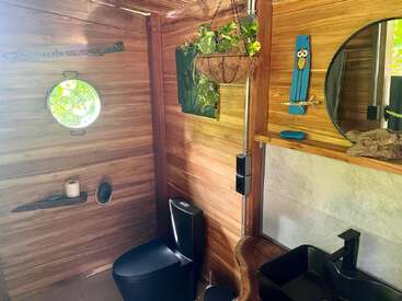 Cette salle de bains confortable se caractérise par des murs en bois, une fenêtre ronde, des toilettes et un lavabo noirs, une plante suspendue, un miroir rond, une décoration en forme de hibou et une lumière naturelle créant une ambiance rustique.