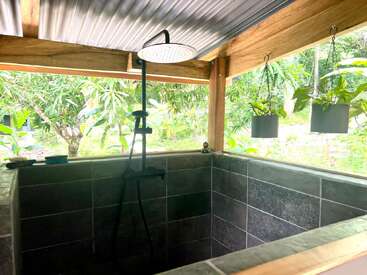 Cette image montre une douche extérieure avec un pommeau de douche noir, des murs carrelés gris, des plantes en pot suspendues et un jardin verdoyant visible à travers des baies vitrées.