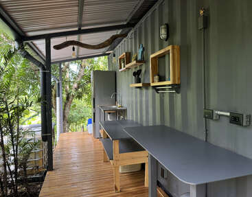 Il s'agit d'un espace de travail ou d'une cuisine en plein air avec des sols en bois, des comptoirs gris, des étagères, un petit évier, un réfrigérateur et un décor moderne, le tout entouré d'une végétation luxuriante.