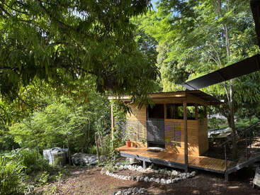 Une petite cabane en bois est nichée dans une forêt verdoyante. La lumière du soleil filtre à travers les arbres, créant une atmosphère sereine et paisible. Allée et plante en pot visibles.