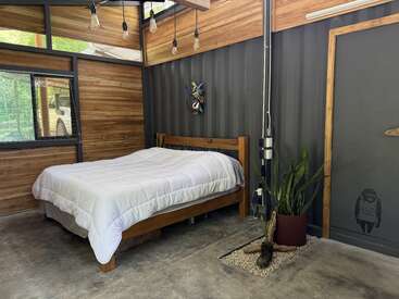 Cette chambre confortable comprend un lit en bois avec une literie blanche, un éclairage moderne, des plantes en pot, des murs en bois, de grandes fenêtres et des touches industrielles, créant ainsi une atmosphère sereine.