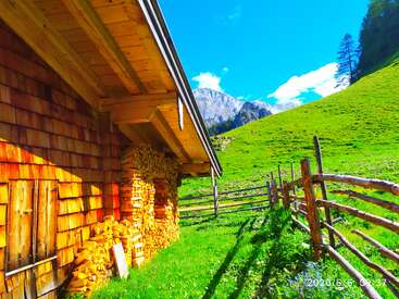 Una rústica cabaña de madera se asienta sobre una ladera de un verde vibrante. La leña apilada, las vallas de madera, la luz del sol y el paisaje montañoso completan esta apacible y pintoresca escena paisajística.