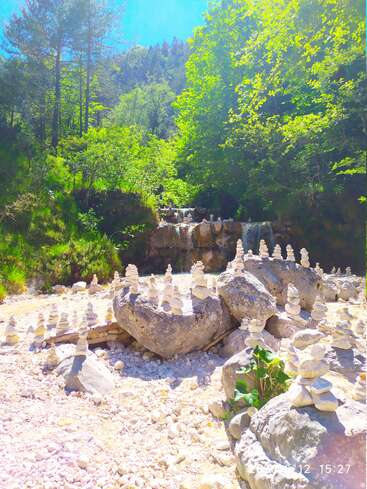 La brillante luz del sol ilumina una tranquila escena forestal con mojones de piedra apilados, el lecho rocoso de un arroyo, frondosos árboles verdes y una pequeña cascada al fondo.