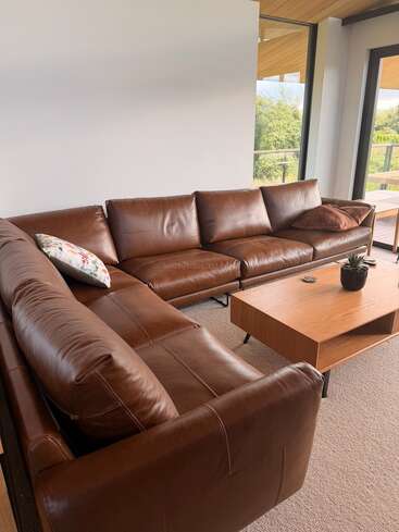 Una sala de estar moderna cuenta con un gran sofá modular de cuero marrón, una mesa de centro de madera, cojines decorativos, una planta en maceta, paredes de colores neutros y grandes ventanas con vistas al exterior.