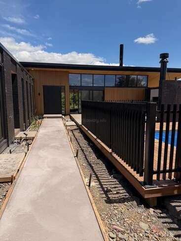 A modern house with large glass windows, wooden panel exterior, and black accents. Concrete pathway leads to the entrance, beside a fenced pool area, under blue skies.