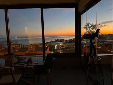 La imagen representa una habitación serena con un telescopio, con vistas a un paisaje urbano y al océano al atardecer, a través de grandes ventanales que ofrecen una vista impresionante.