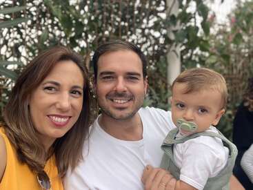 La imagen muestra a una familia de tres miembros posando para una foto, con la madre a la izquierda, el padre en el centro y el bebé a la derecha.