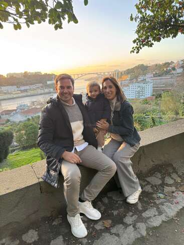 The image depicts a family of three posing for a photo on a stone wall, with a scenic view of a bridge and cityscape in the background.