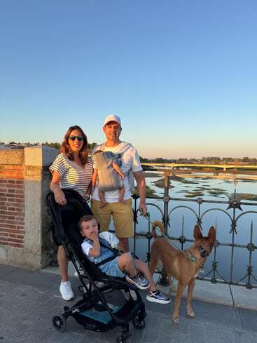 Una familia posa en un paseo junto al río al atardecer. Una mujer, un hombre con un bebé en brazos, un niño en cochecito y un perro marrón disfrutan juntos del paisaje.