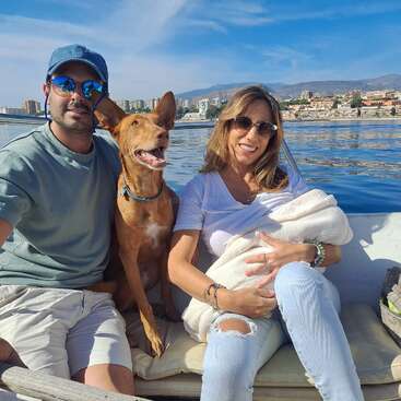 La imagen muestra a un hombre, una mujer y un perro sentados en un barco, con el perfil de la ciudad y el agua de fondo, en un día soleado de cielo azul.