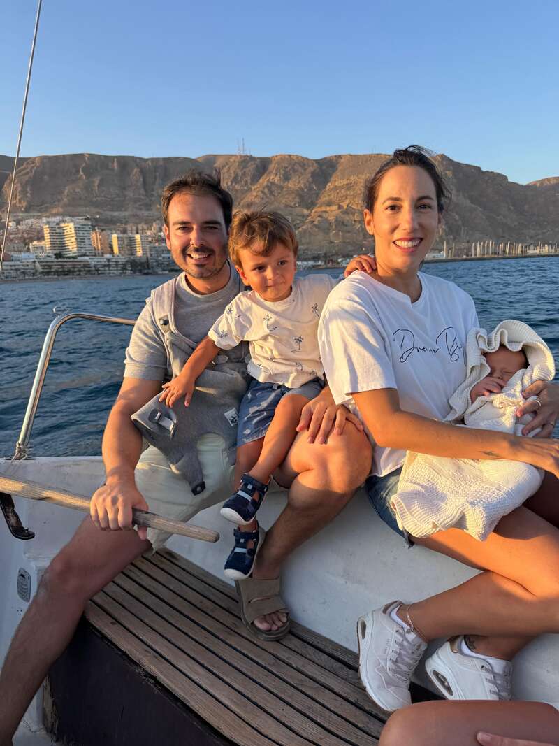 Una feliz familia de cuatro miembros sentados juntos en un barco, sonriendo con las montañas y una ciudad como telón de fondo, disfrutando de un día soleado en el agua.