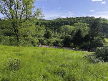 Um prado verde exuberante desce até uma área de jardim cercada. As árvores cobrem as colinas ao fundo sob um céu claro e levemente nublado.