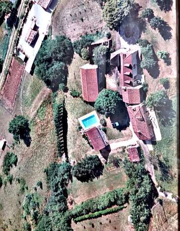 Esta imagem aérea mostra uma propriedade rural com vários edifícios de telhado vermelho, uma piscina, campos verdes, jardins, árvores, entradas de garagem e um carro estacionado.