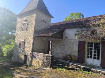 Esta imagem mostra uma antiga casa de pedra com uma torre pontiaguda, persianas rústicas de madeira, uma pequena varanda, cercada por vegetação, banhada pela luz do sol.