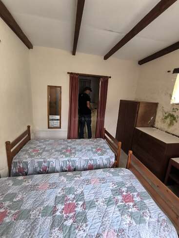 Um quarto pequeno e simples com duas camas de solteiro, colchas florais, móveis de madeira, um espelho e uma pessoa em pé perto de uma porta com cortinas. As paredes apresentam algum desgaste.