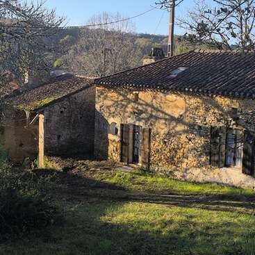 A rustic stone cottage with wooden shutters sits in a grassy yard, surrounded by trees and bathed in warm, late afternoon sunlight in the countryside.
