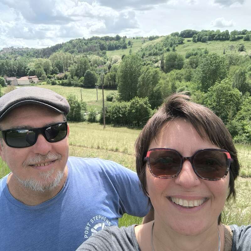 Um casal sorridente usando óculos escuros tira uma selfie em um campo verde e exuberante, com colinas, árvores e céu nublado ao fundo. Atmosfera pacífica e feliz.