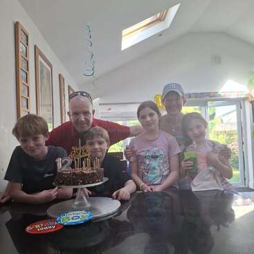 Une famille célèbre le 12e anniversaire d'un garçon avec un gâteau au chocolat, des bougies, des décorations et des sourires, réunis dans une cuisine lumineuse et joyeuse.