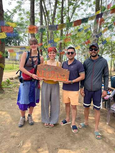 Four people stand outdoors, holding a colorful "NIRVANA" sign. They are surrounded by trees and vibrant prayer flags, smiling and dressed in casual, comfortable clothing.